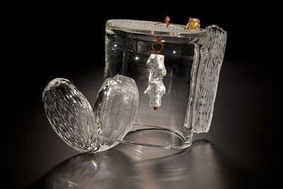 Glass sculpture with a cylindrical base, two wing-like shapes, and a hanging, textured object.