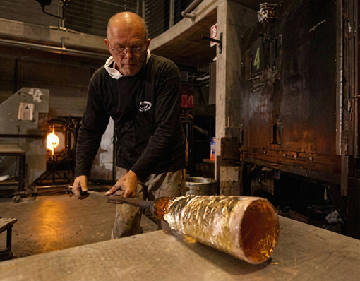 Man shaping molten glass with tools in a workshop.
