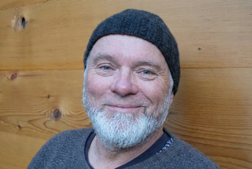 Man with white beard, dark beanie, and grey sweater smiles against a wood-paneled wall.