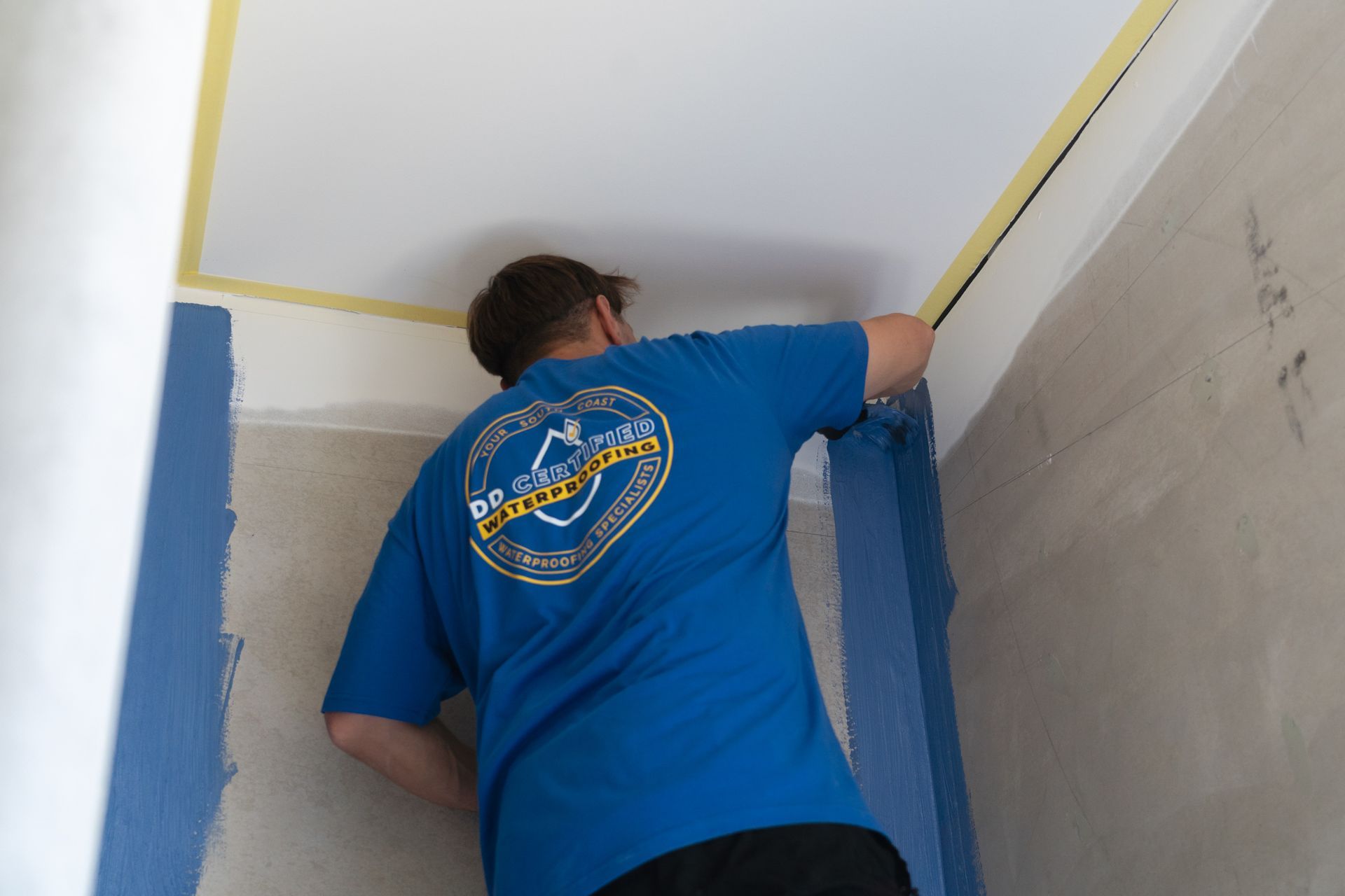 A man wearing a blue shirt with a logo on the back is painting a wall
