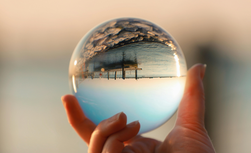 Hand holding crystal ball, reflecting a waterfront view with clouds and pier.