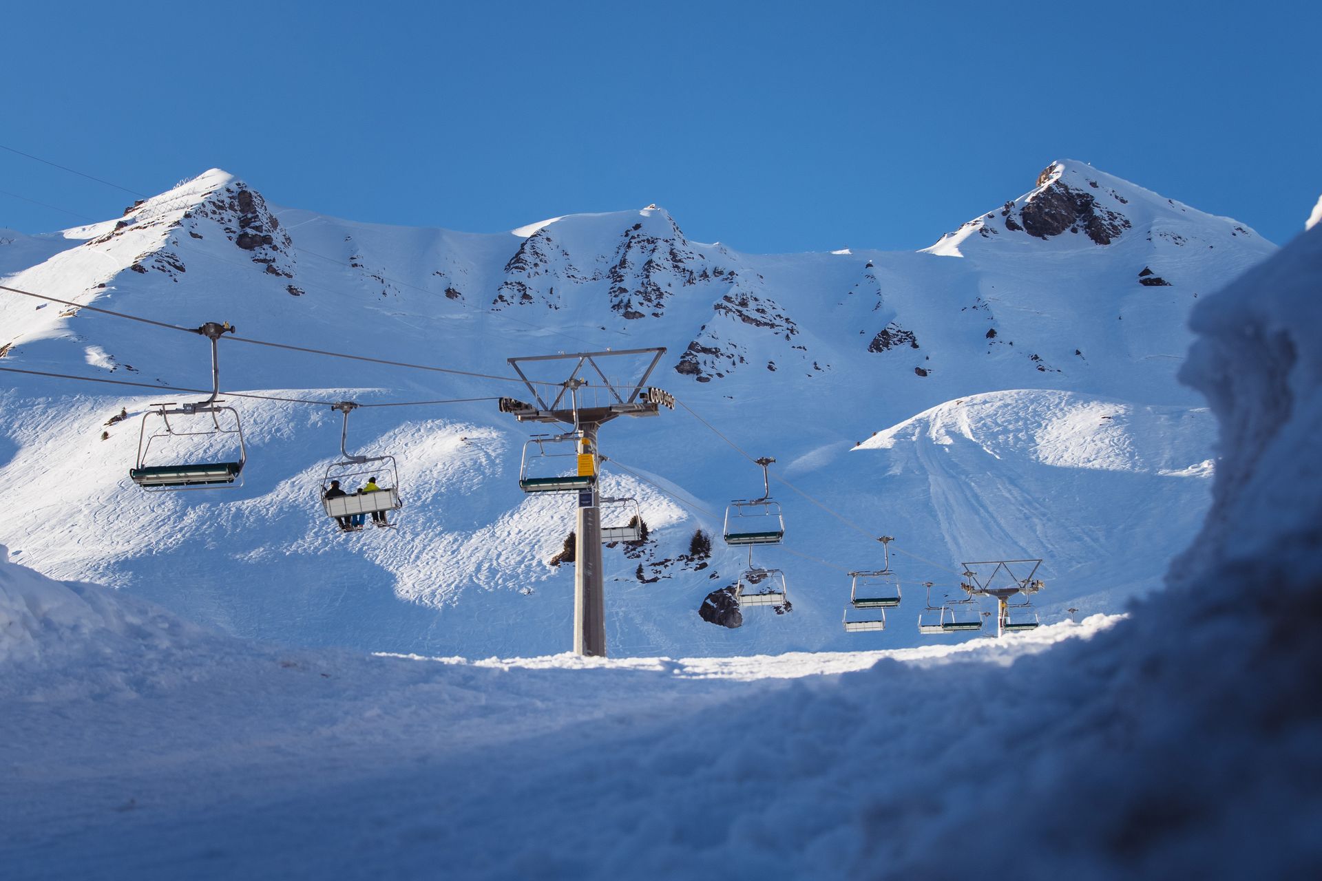 Ski lift with skiers ascending snow-covered mountain peaks under a clear blue sky.