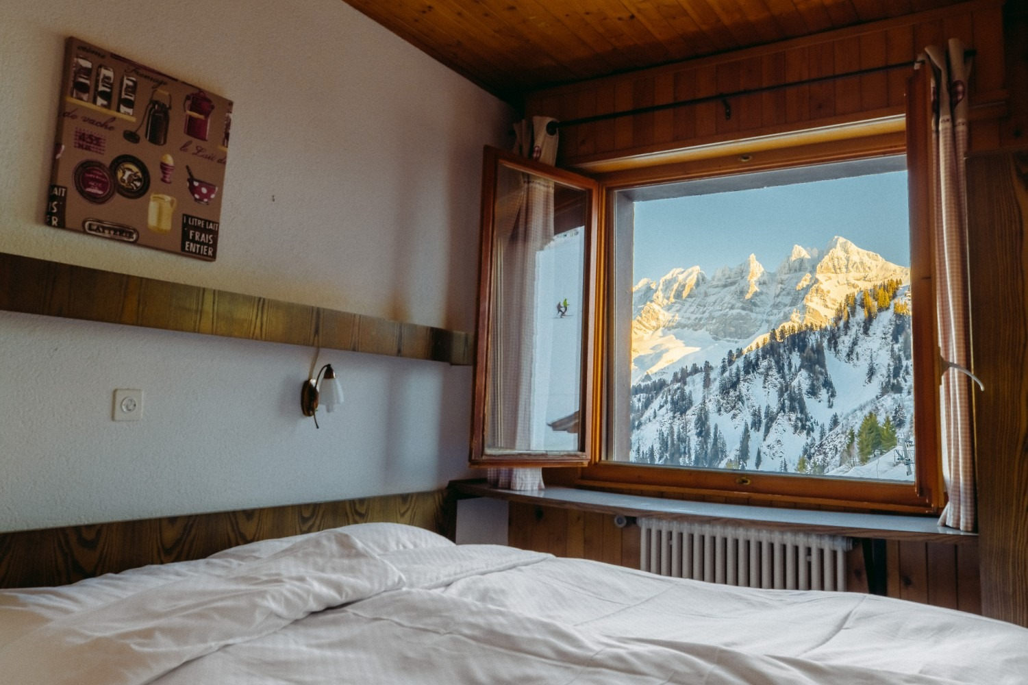 Cozy bedroom with a mountain view framed by a wooden window. Snow-capped peaks are visible.