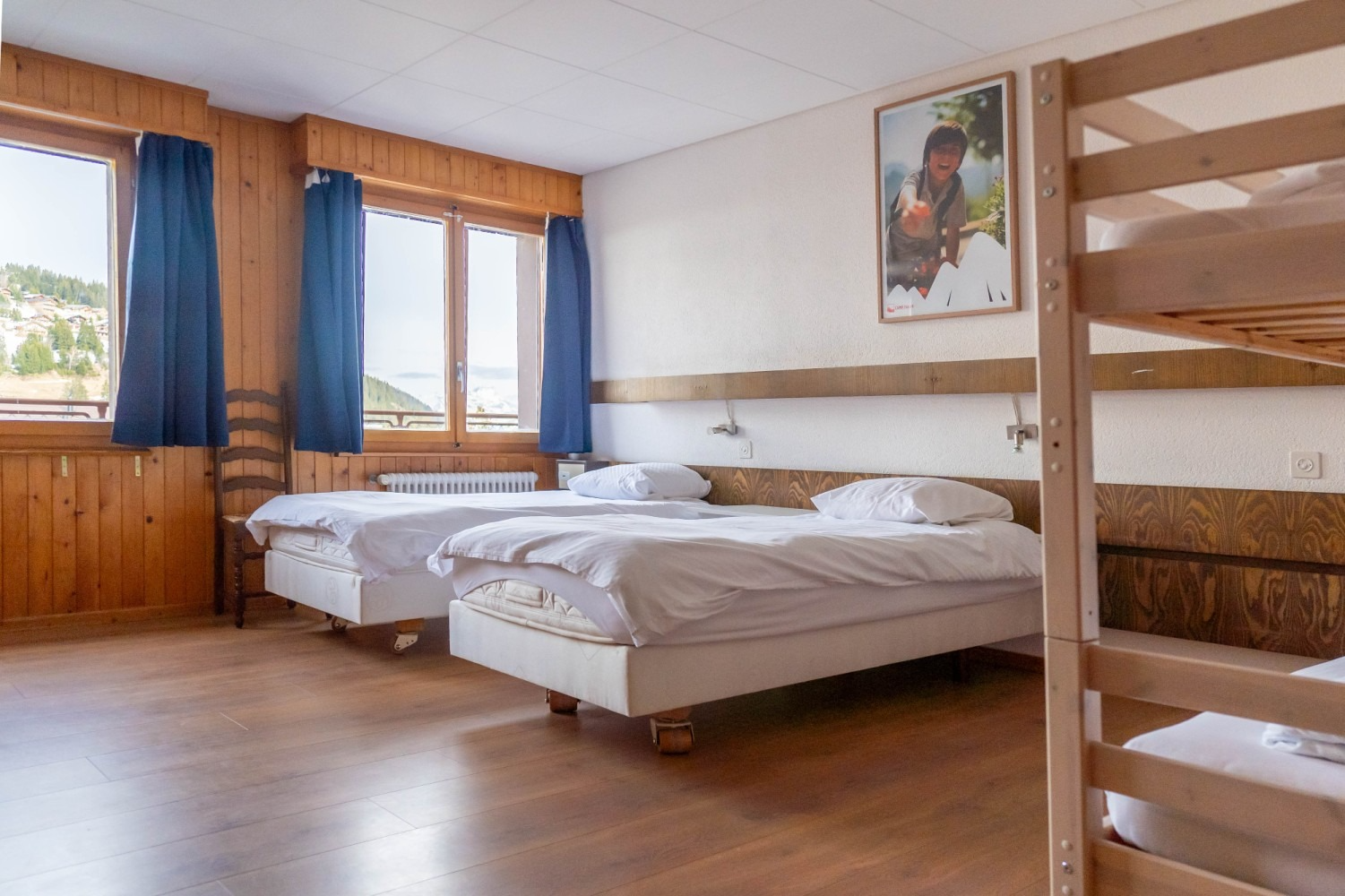 A brightly lit hostel room with two single beds, a bunk bed, and two windows with blue curtains.