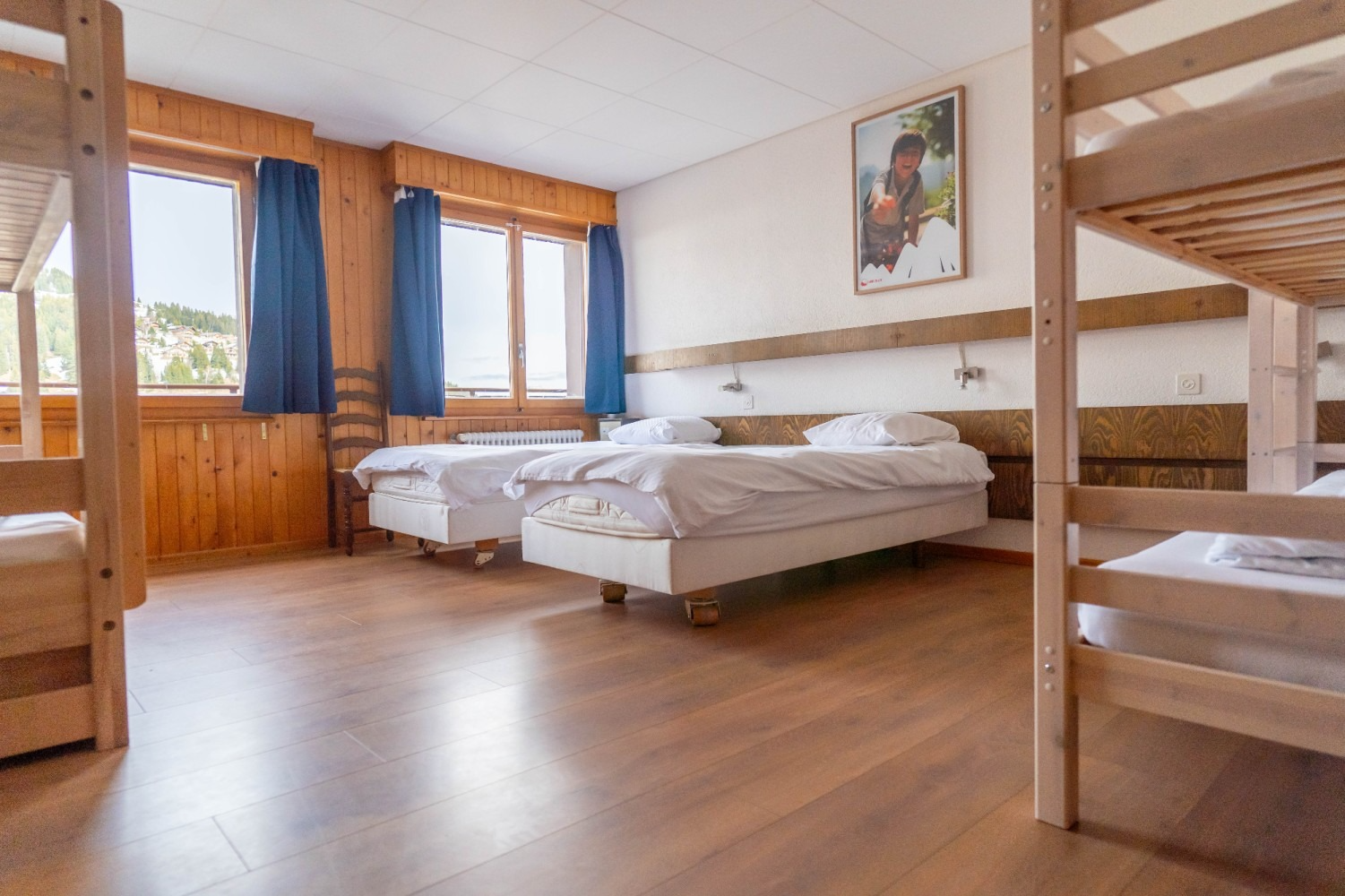 A dorm room with two beds, a window with blue curtains, and bunk beds.