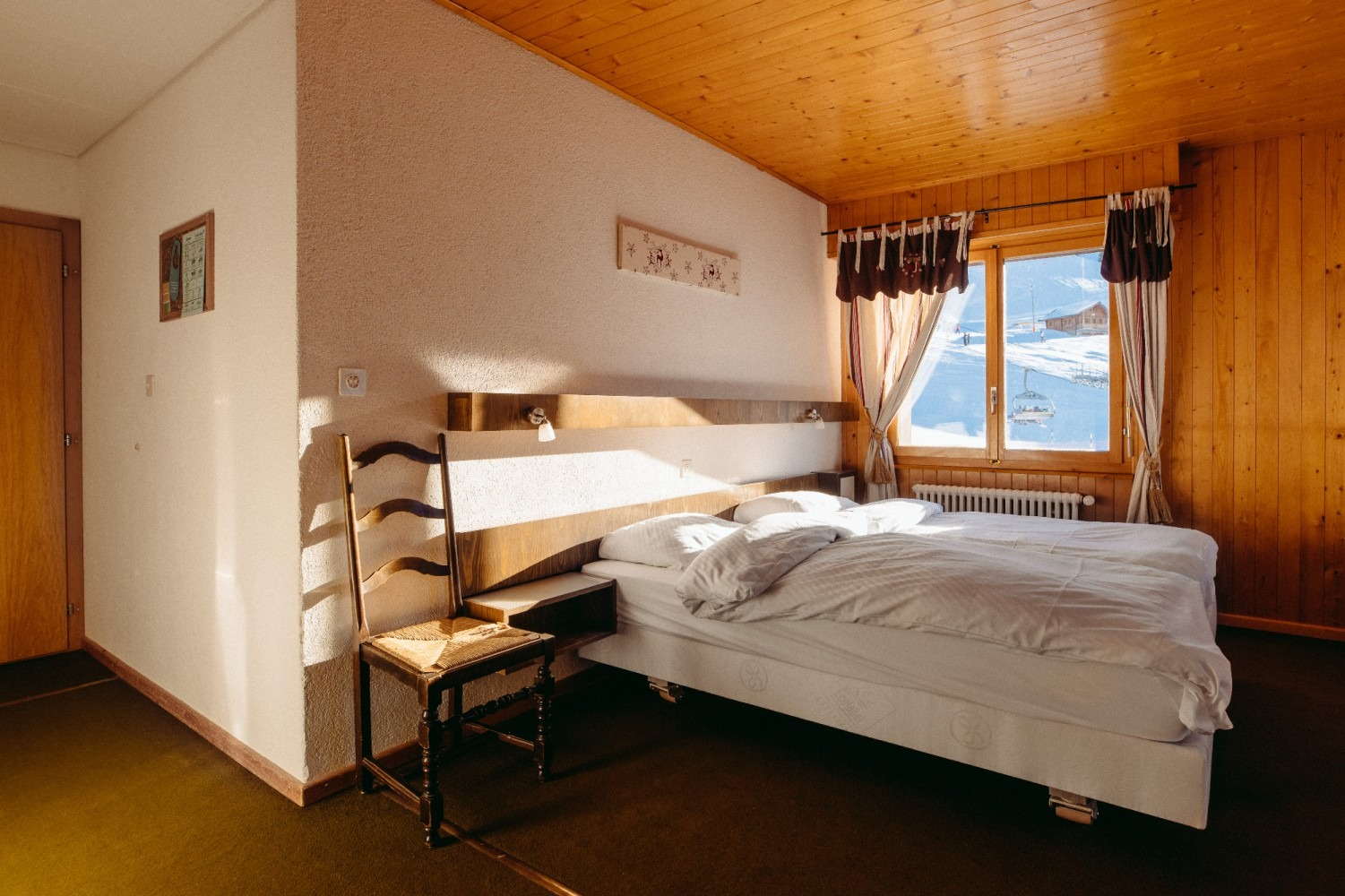 Cozy hotel room with wooden walls, a double bed, and a window overlooking a snowy mountain scene.