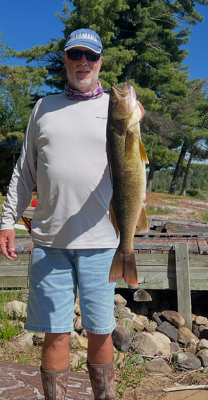 Man holding a large fish outdoors by a lakeside dock with trees in the background