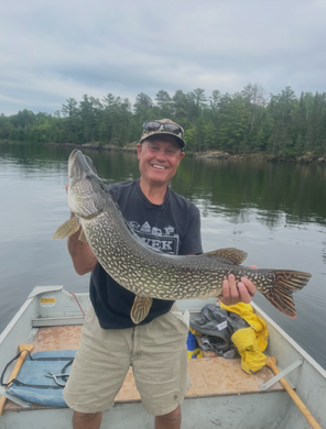 Smiling man on a boat holding a large fish on a calm lake, with forested shore in the background