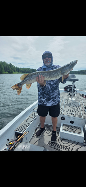 Person on a boat holding a large fish over the water, wearing a hooded jacket and shorts.