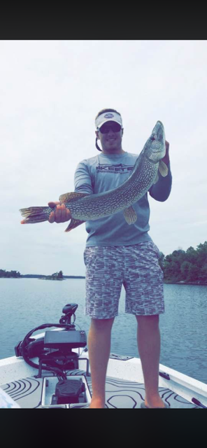 Person on a boat holding a large fish over the water, wearing a cap and patterned shorts.