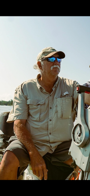 Man in beige shirt and cap sitting on a boat, wearing sunglasses under a bright sky.