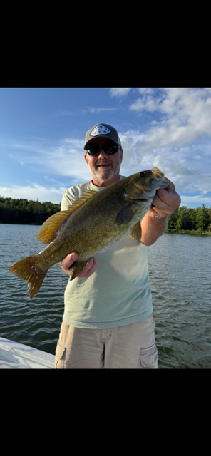 Person holding a large fish on a lake, smiling under a blue sky.