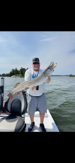 Person standing on a boat holding a large fish on a sunny lake.