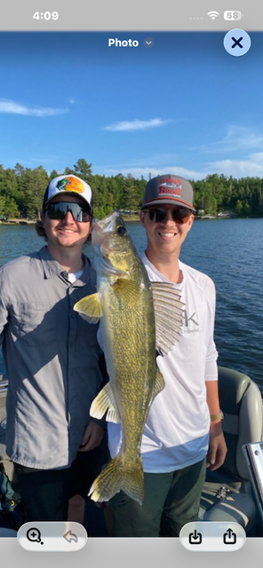 Two people holding a large fish on a lake, both wearing caps and smiling in a boat
