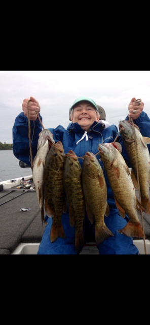 Person in a blue jacket holding up several large fish on a boat, smiling on a cloudy day.