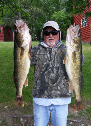 Person in camo vest and cap holds up two large fish outdoors on a grassy path.