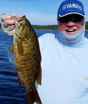 Man smiling and holding a large fish on a boat, with a blue Yamaha cap and bright sky behind him