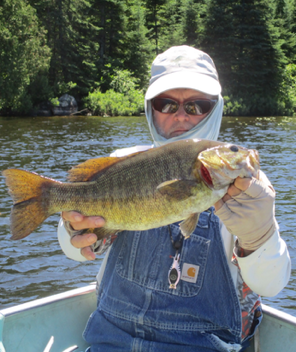 Person holding a large fish on a riverbank, wearing sunglasses and a cap, with water and trees behind.