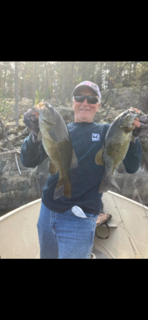 Man in a boat holding up two large fish, wearing sunglasses and a cap in a wooded setting