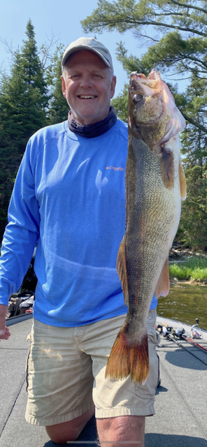 Man holding a large fish outdoors on a sunny dock, wearing a blue shirt and cap.