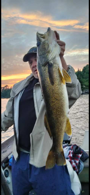 Person holding a large fish by the water at sunset, with cloudy sky and boat in background