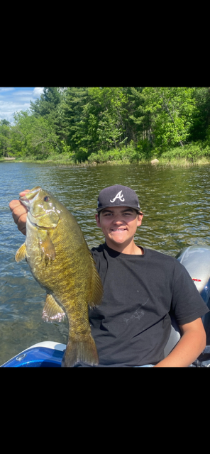 Person in a black shirt and cap holds a large fish while sitting in a boat on a lake.