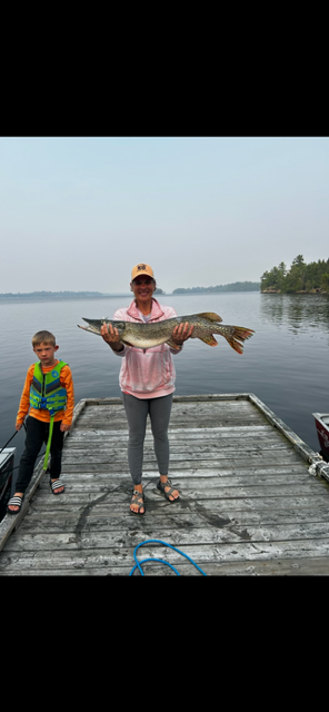 Person holding a large fish on a dock beside a child, with calm water and trees in the background