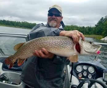 Smiling angler holding a large northern pike on a boat, with cloudy lake and trees in the background