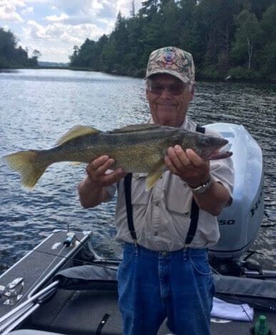 Man holding a large fish on a boat on a lake, with trees and cloudy sky in the background