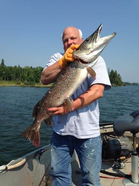 Man holding a large fish on a boat, with a lake and treeline in the background.