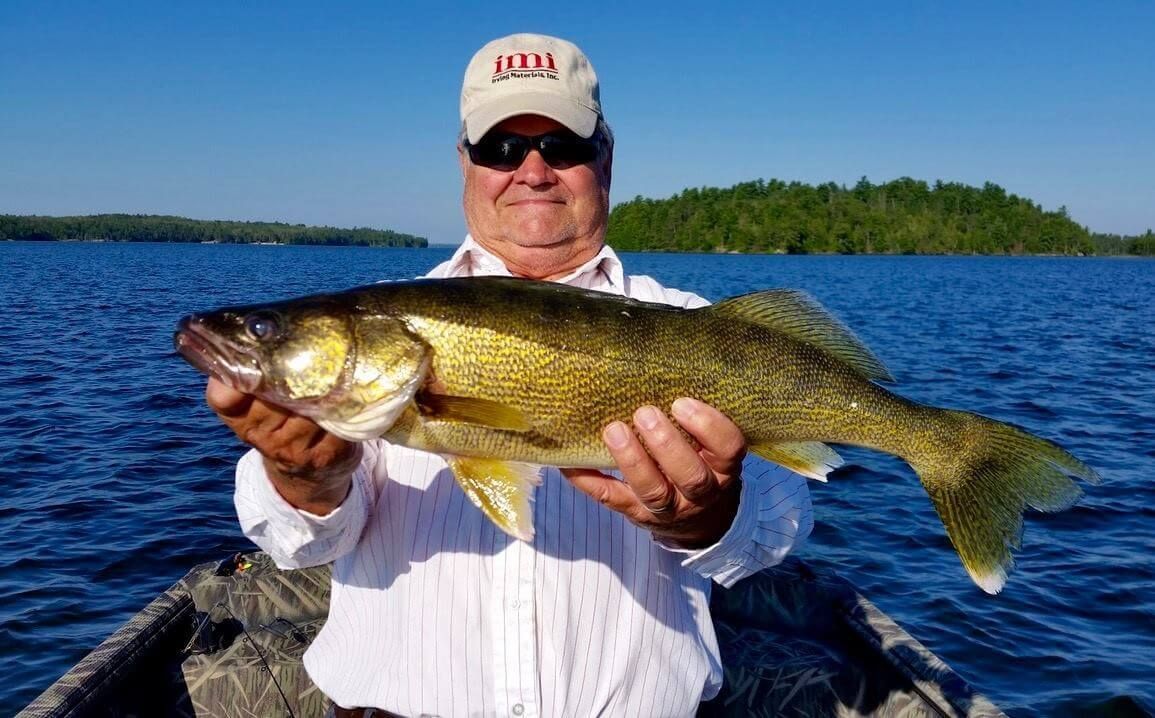 Man holding a large fish on a boat, with blue water and a tree-lined shoreline behind him.