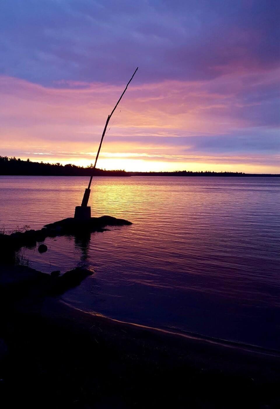 Silhouette of a fishing rod on a lakeshore at sunset, with purple and orange sky reflecting on the water