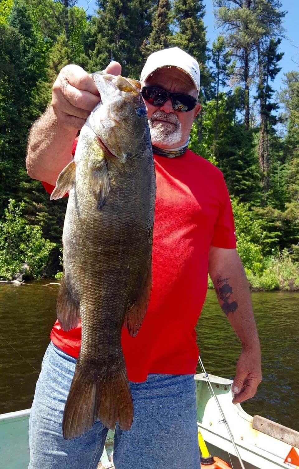 Man in red shirt holding a large fish on a boat beside a forested lake