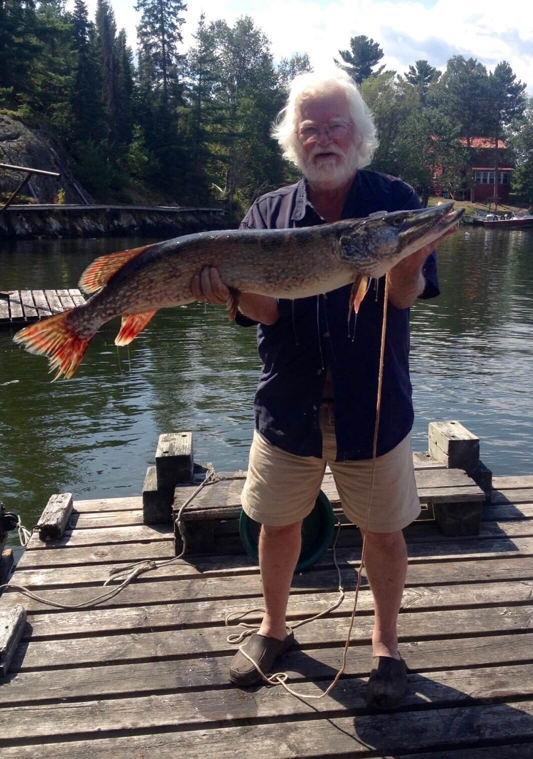 Man holding a large fish on a dock by a lake, with trees and calm water in the background
