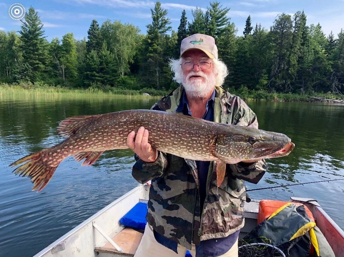 Man holding a large fish on a boat, with calm lake and forest shoreline behind him.