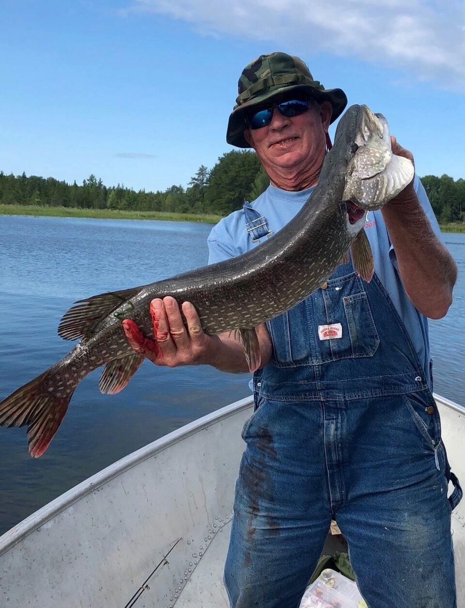 Person in overalls holds a large fish on a boat by a lake under a blue sky.