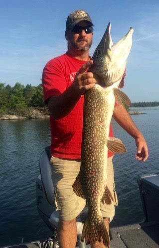 Man in red shirt and khaki shorts holding a large fish on a boat on a calm lake