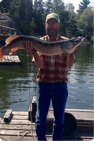 Man holding a large fish on a dock by a lake, with trees and a canoe in the background.