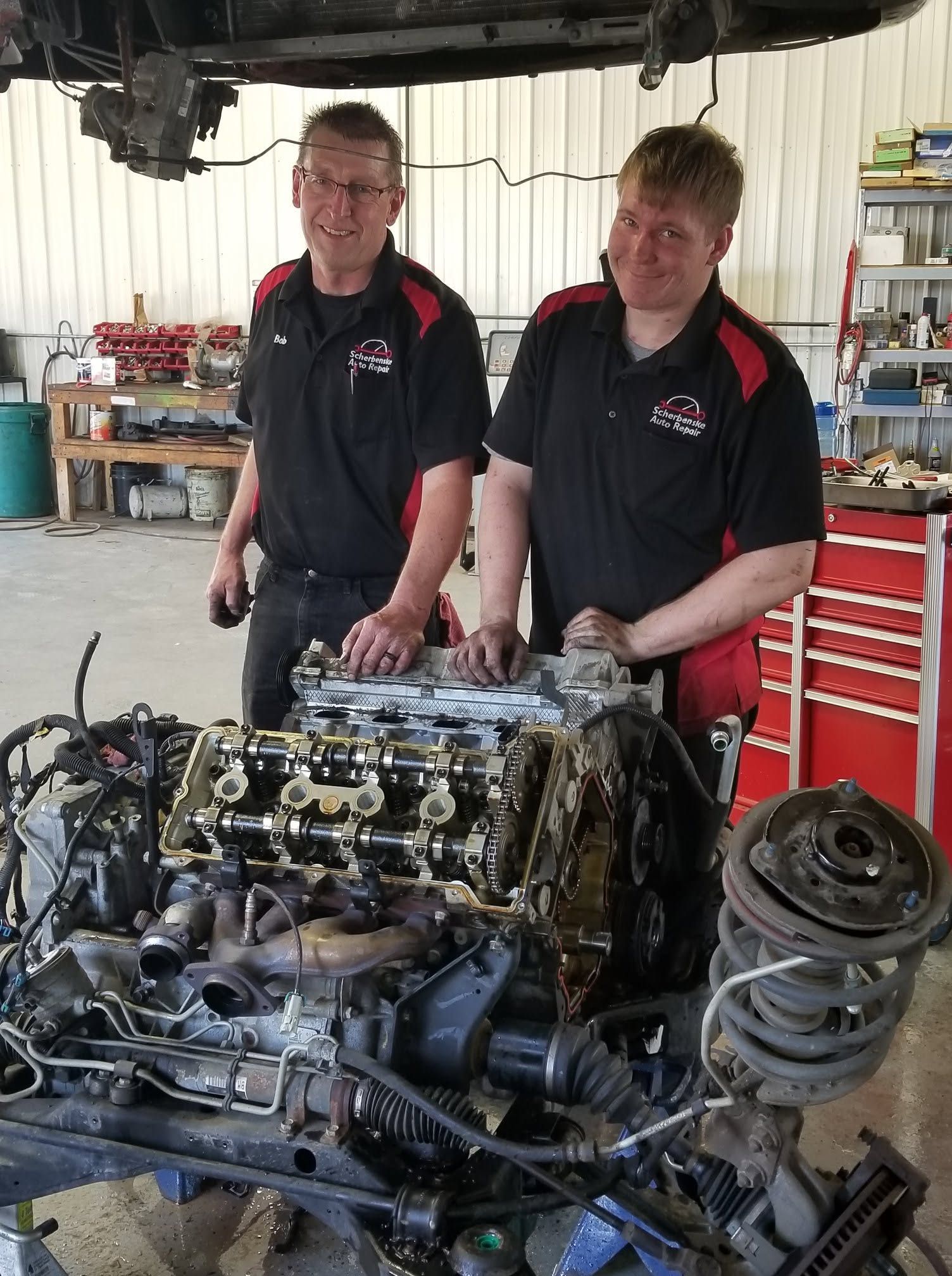 Two mechanics in black shirts stand beside a car engine in a workshop, with tools and parts around them | Scherbenske Auto Repair Inc
