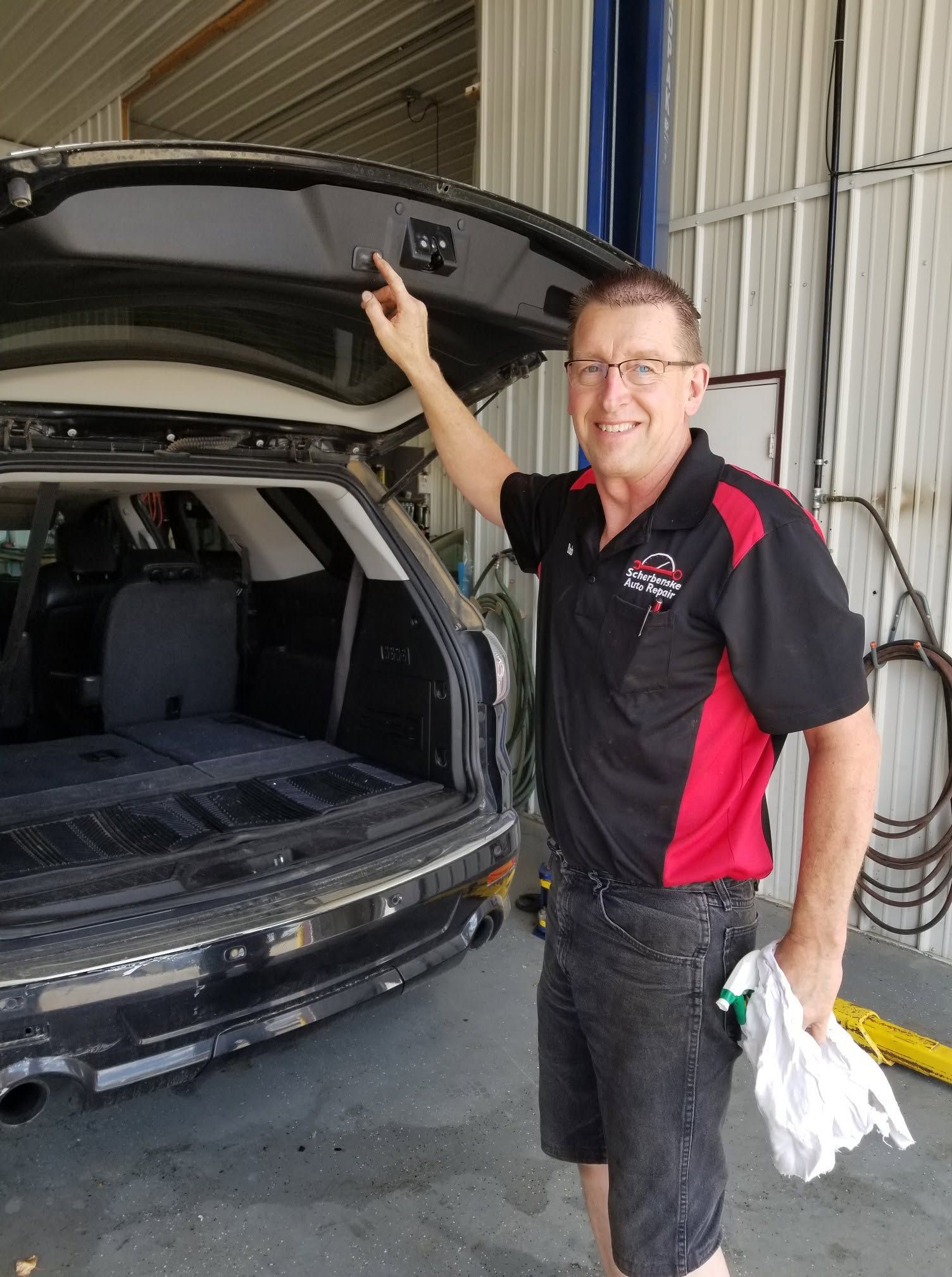 Man in black and red work shirt inspecting an open SUV hatch in a garage | Scherbenske Auto Repair Inc