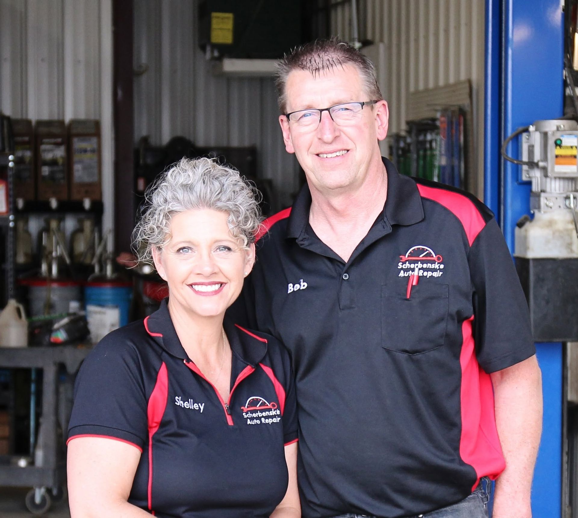 Two mechanics in black-and-red uniforms smiling in a workshop doorway | Scherbenske Auto Repair Inc