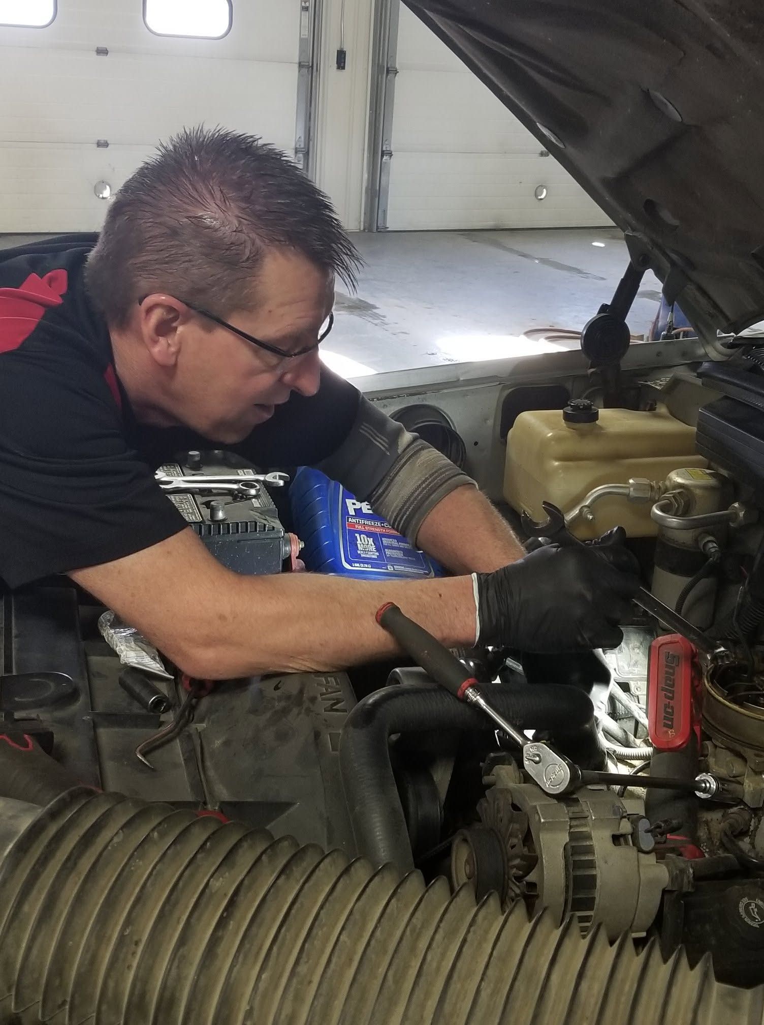 Mechanic working under an open car hood in a garage, using tools on the engine. | Scherbenske Auto Repair Inc