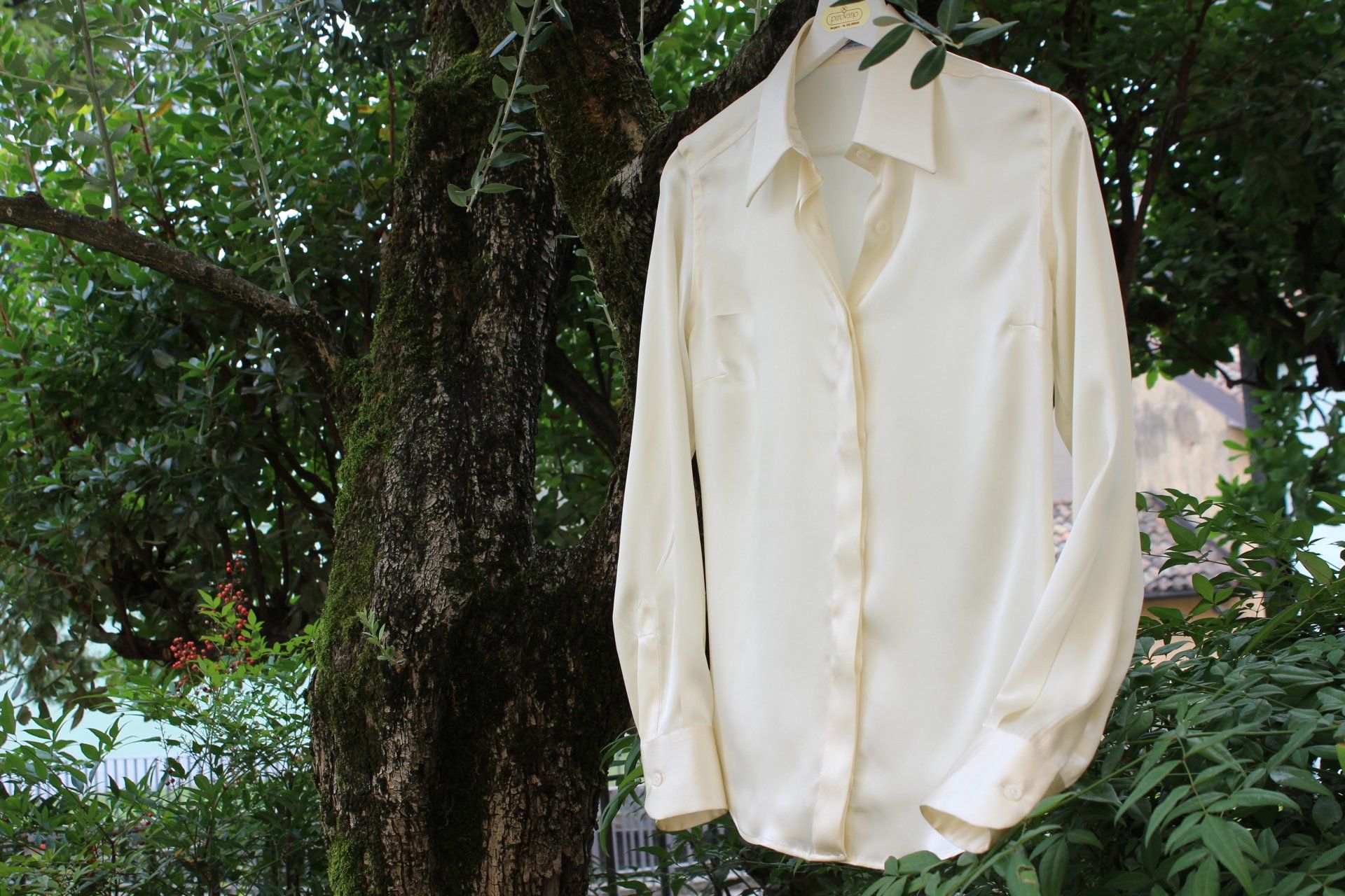 Camicia color crema abbottonata appesa all'aperto tra alberi e vegetazione.