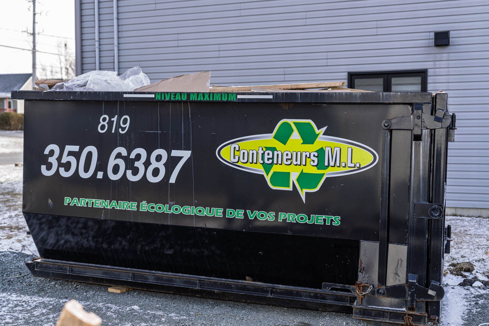 Une remorque benne noire portant l'inscription « 350.6387 » et un logo vert-jaune est stationnée sur un sol enneigé près d'un bâtiment.