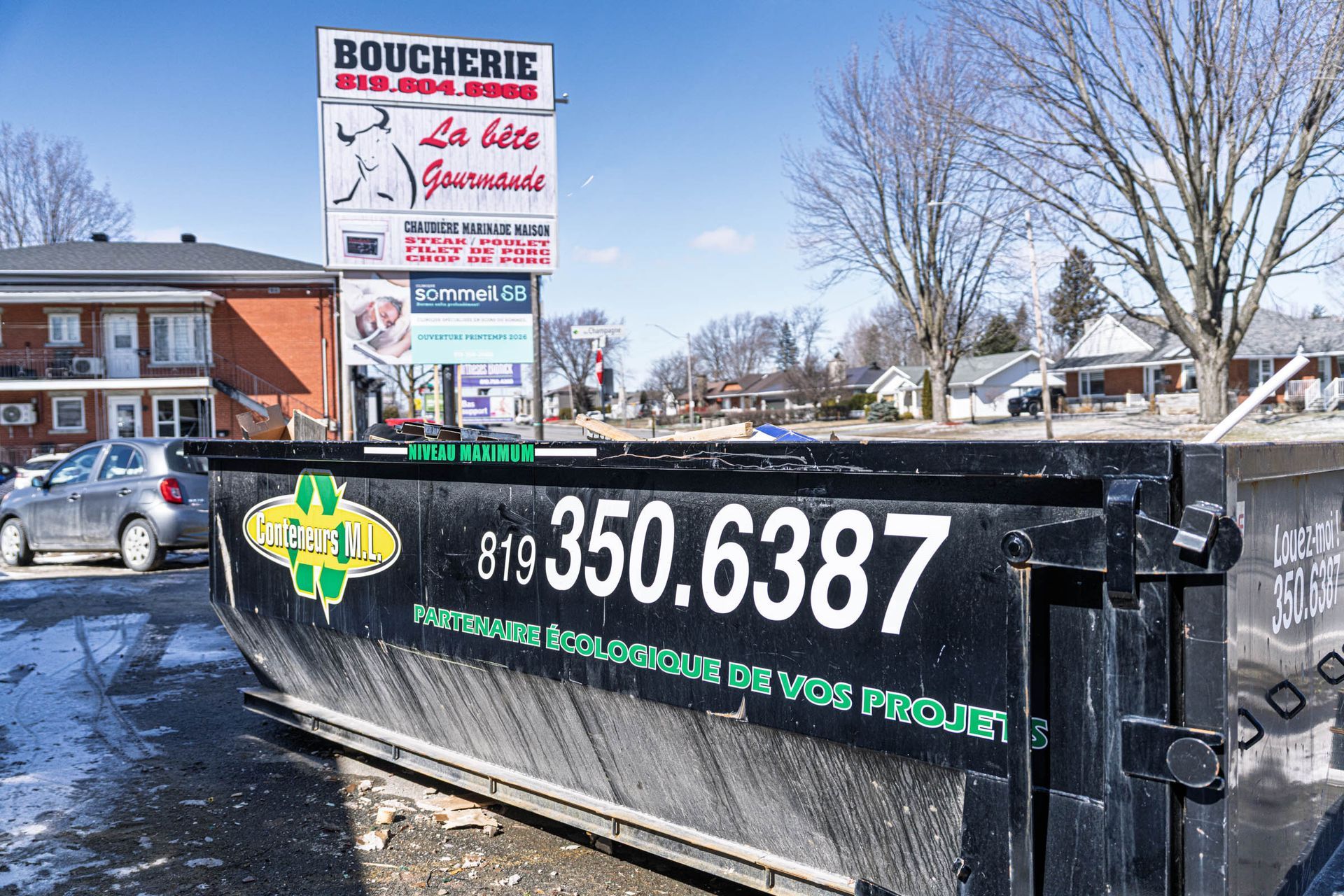 Conteneur à ordures noir portant le numéro 350-6387 sur un parking enneigé près d'une enseigne de boucherie