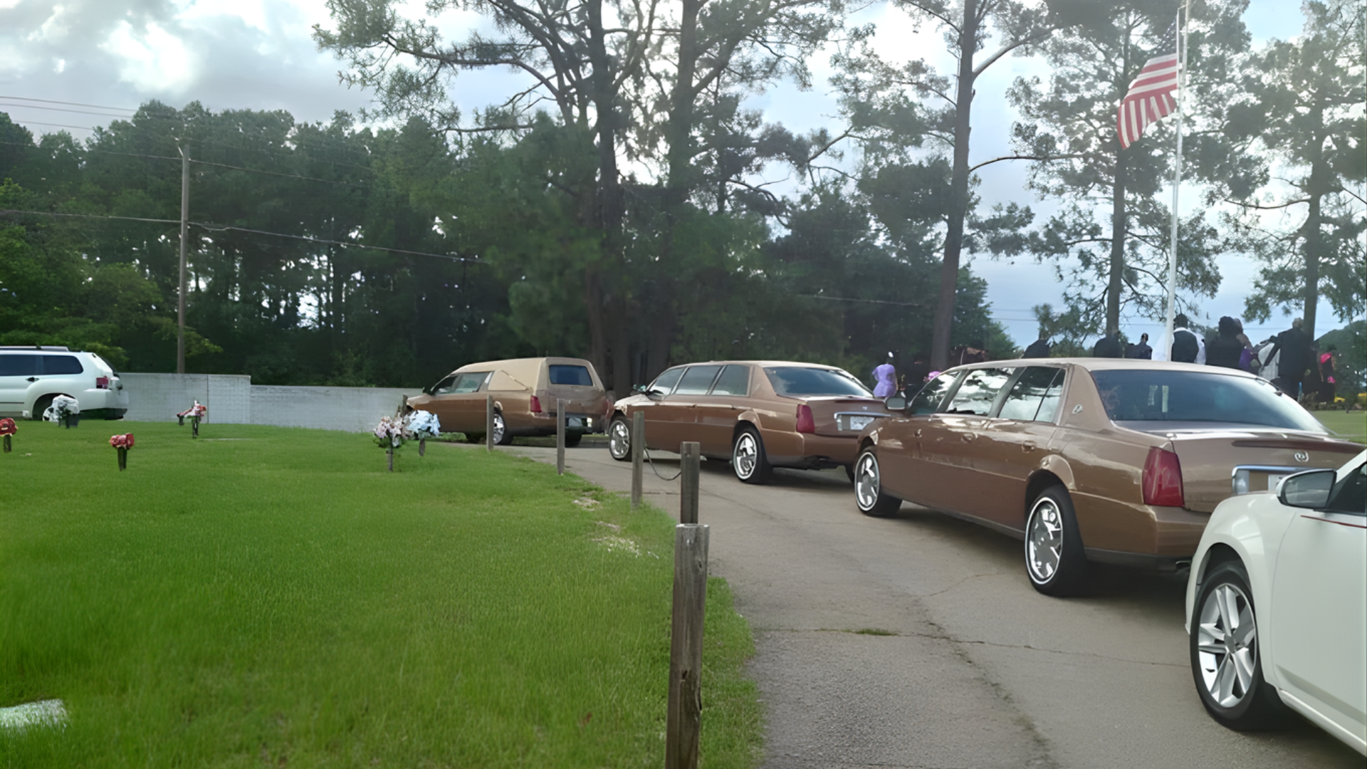 A funeral procession in a cemetery with brown cars and an American flag.