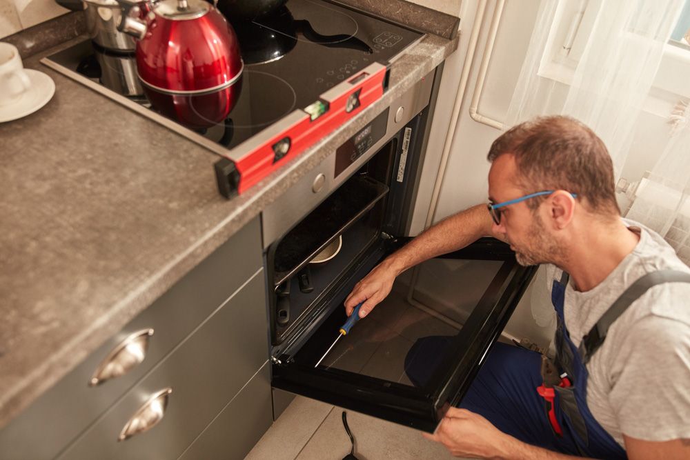 A person in work coveralls uses a screwdriver to repair an oven door while a spirit level rests on the stovetop.
