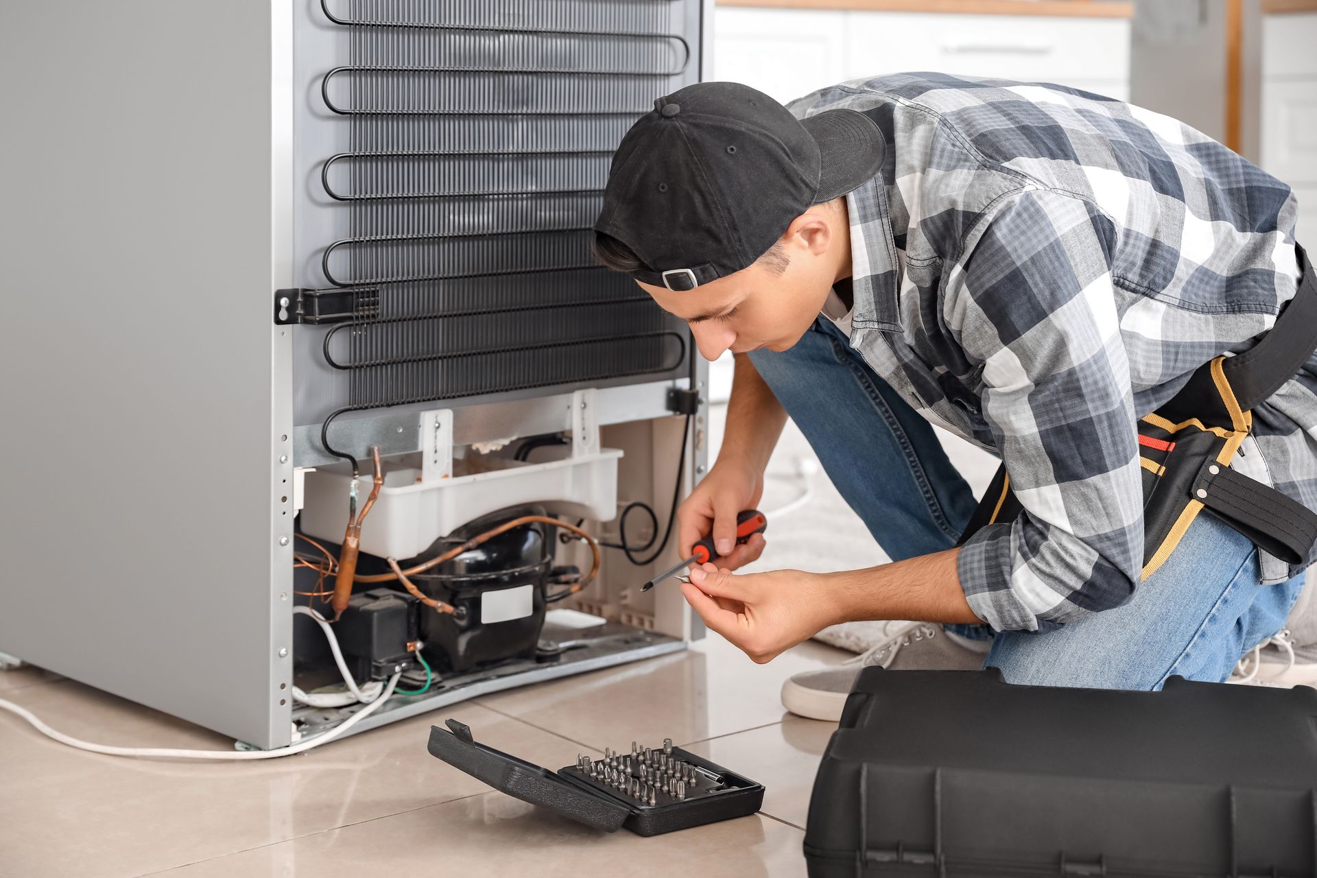 A technician kneeling to repair the compressor and coils on the back of a refrigerator with a screwdriver and tool kit.