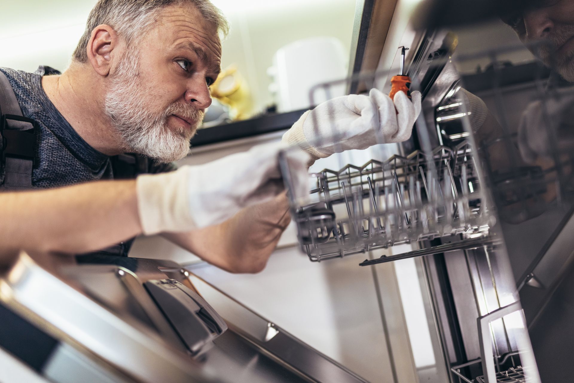A technician in white gloves uses a screwdriver to repair the interior mechanism of an open dishwasher.