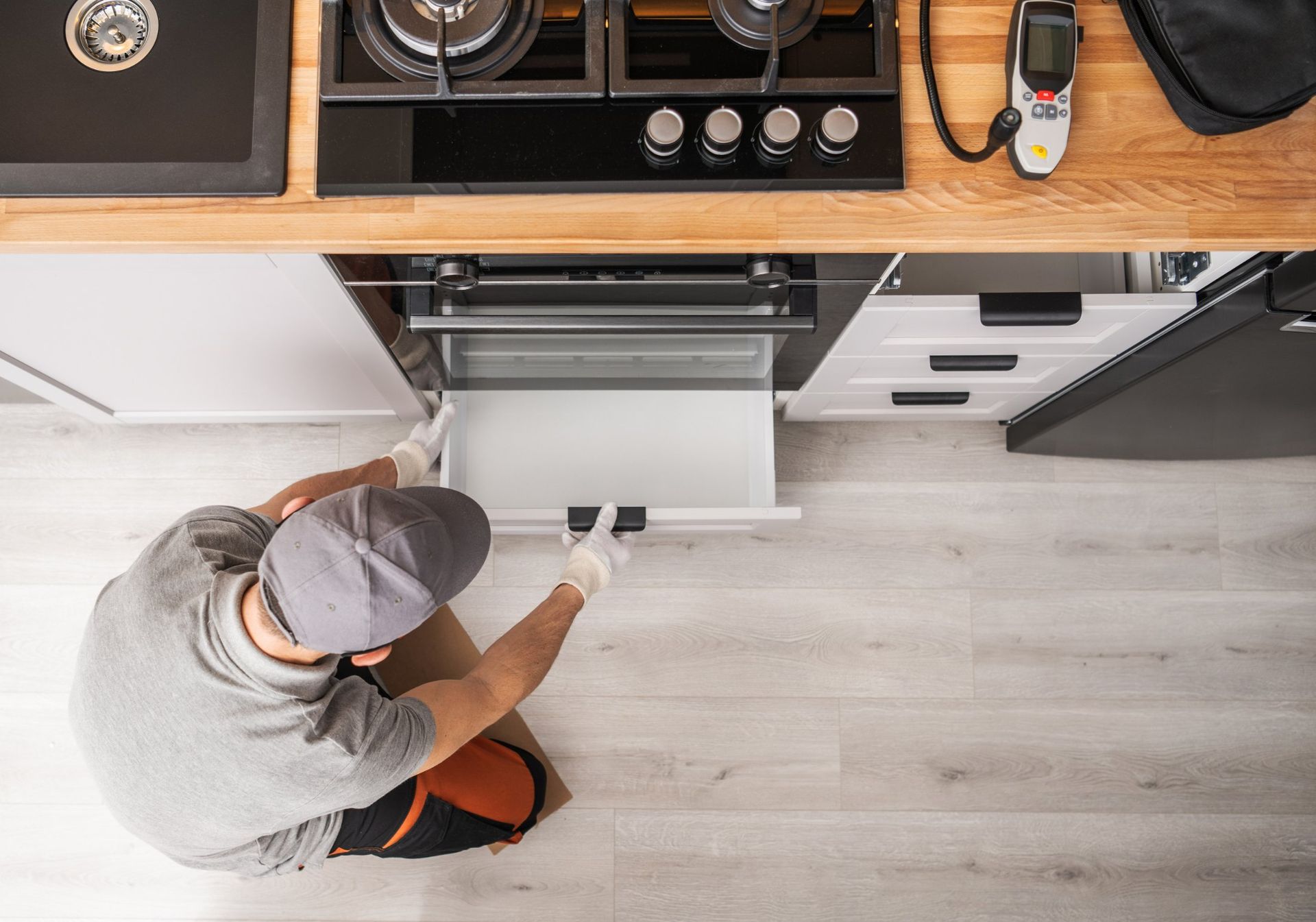 A technician in a grey uniform and gloves pulls open a kitchen oven drawer in a modern kitchen.